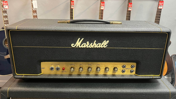 A Marshall 1971 Lead Head JMP 1987 50 Watt amplifier with gold control panel sits atop a speaker cabinet; guitars hang behind, evoking classic British grind. Includes flight case.