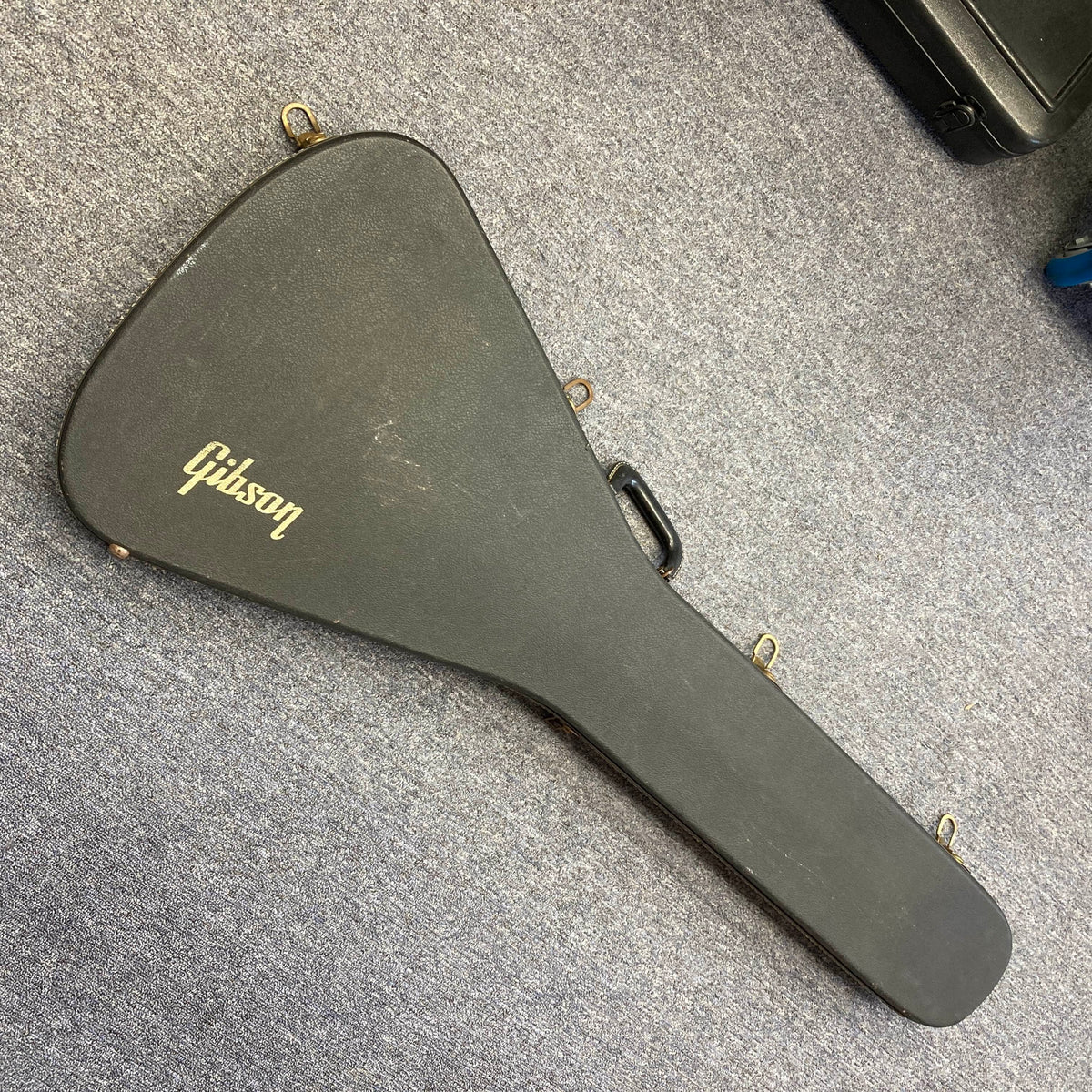 A black, hard-shell Gibson case featuring a gold Gibson logo—designed to fit the 1975 Gibson Flying V w/Case or other vintage Flying V models—is lying closed on a gray carpeted floor.