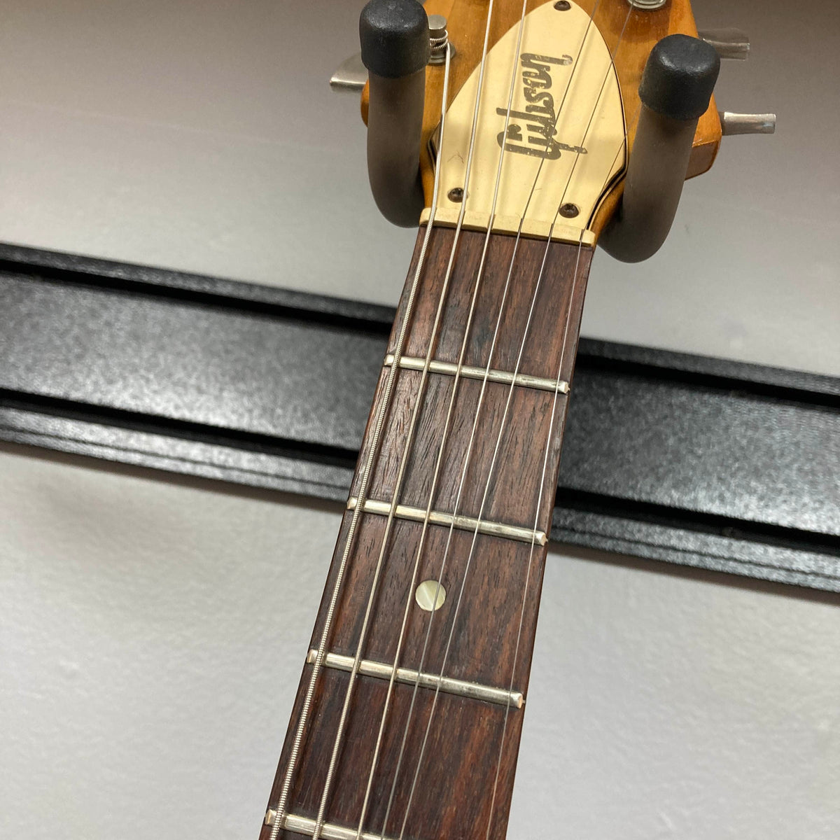 Close-up of the headstock of a 1975 Gibson Flying V w/Case by Gibson, showing part of the fretboard with six strings and a wall-mounted guitar hanger, featuring the iconic vintage Flying V design with a natural finish.