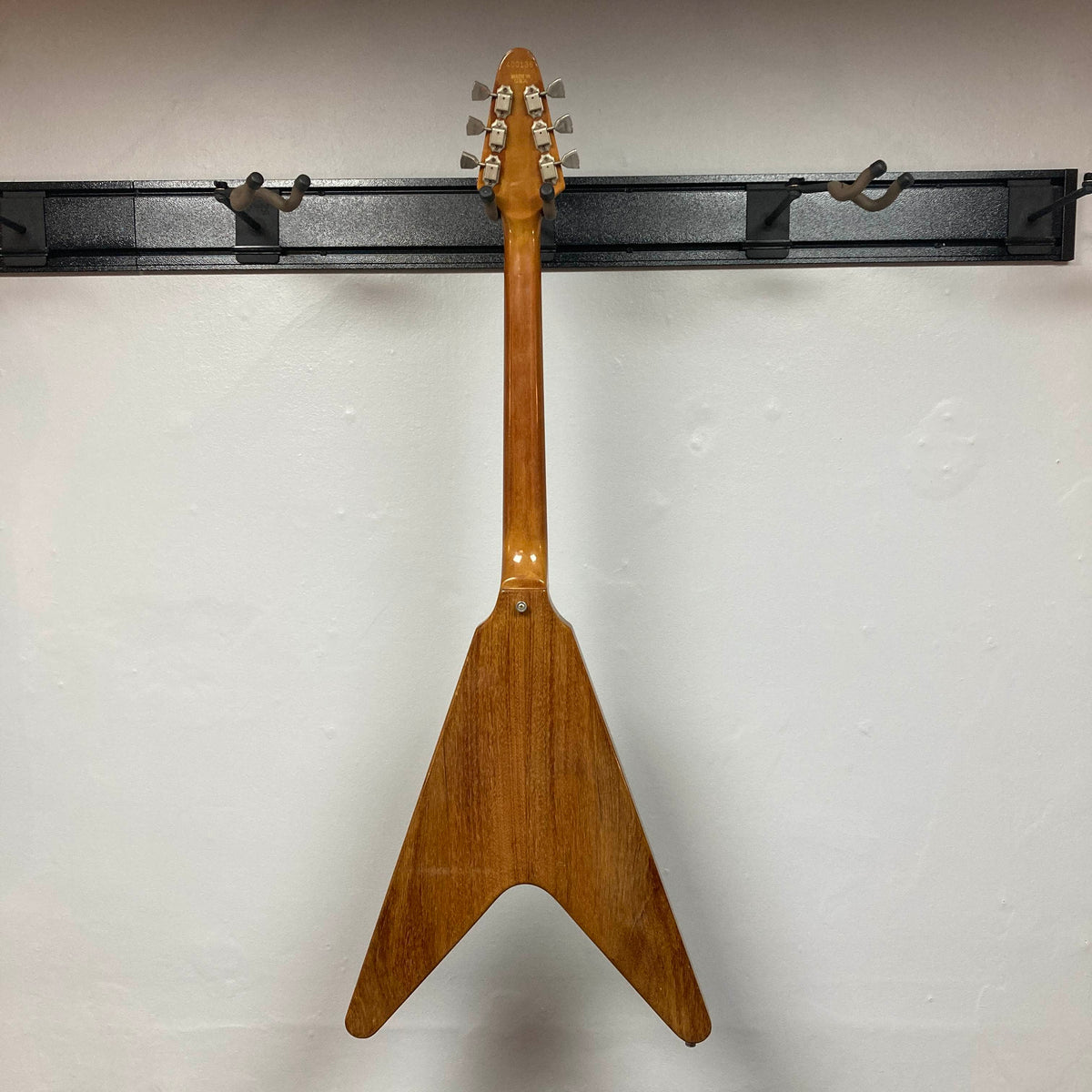 A 1975 Gibson Flying V w/Case by Gibson, featuring a natural finish, hangs by its headstock on a black wall-mounted rack, displayed from the back against a white wall.