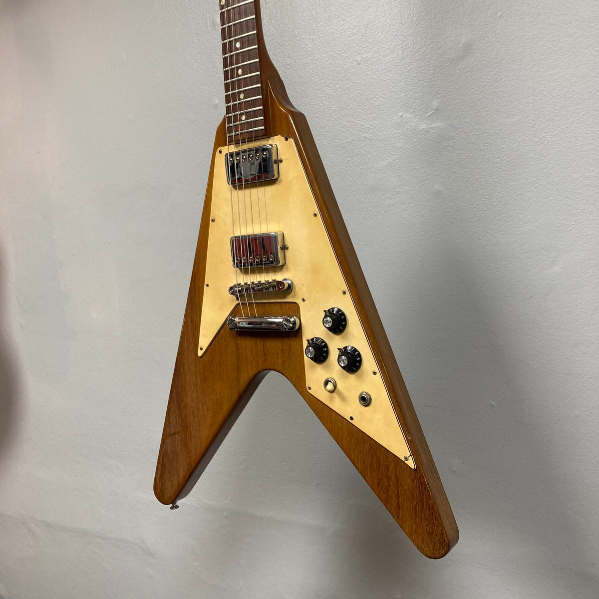 A 1975 Gibson Flying V electric guitar by Gibson, featuring a natural finish, V-shaped body, and cream pickguard, is mounted on a light gray wall.