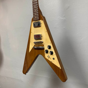 A 1975 Gibson Flying V electric guitar by Gibson, featuring a natural finish, V-shaped body, and cream pickguard, is mounted on a light gray wall.