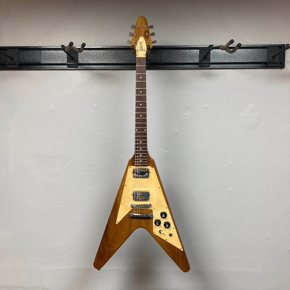 A Gibson 1975 Flying V w/Case, featuring a natural finish, cream pickguard, and dual pickups, is displayed on a black wall-mounted rack against a plain white wall.