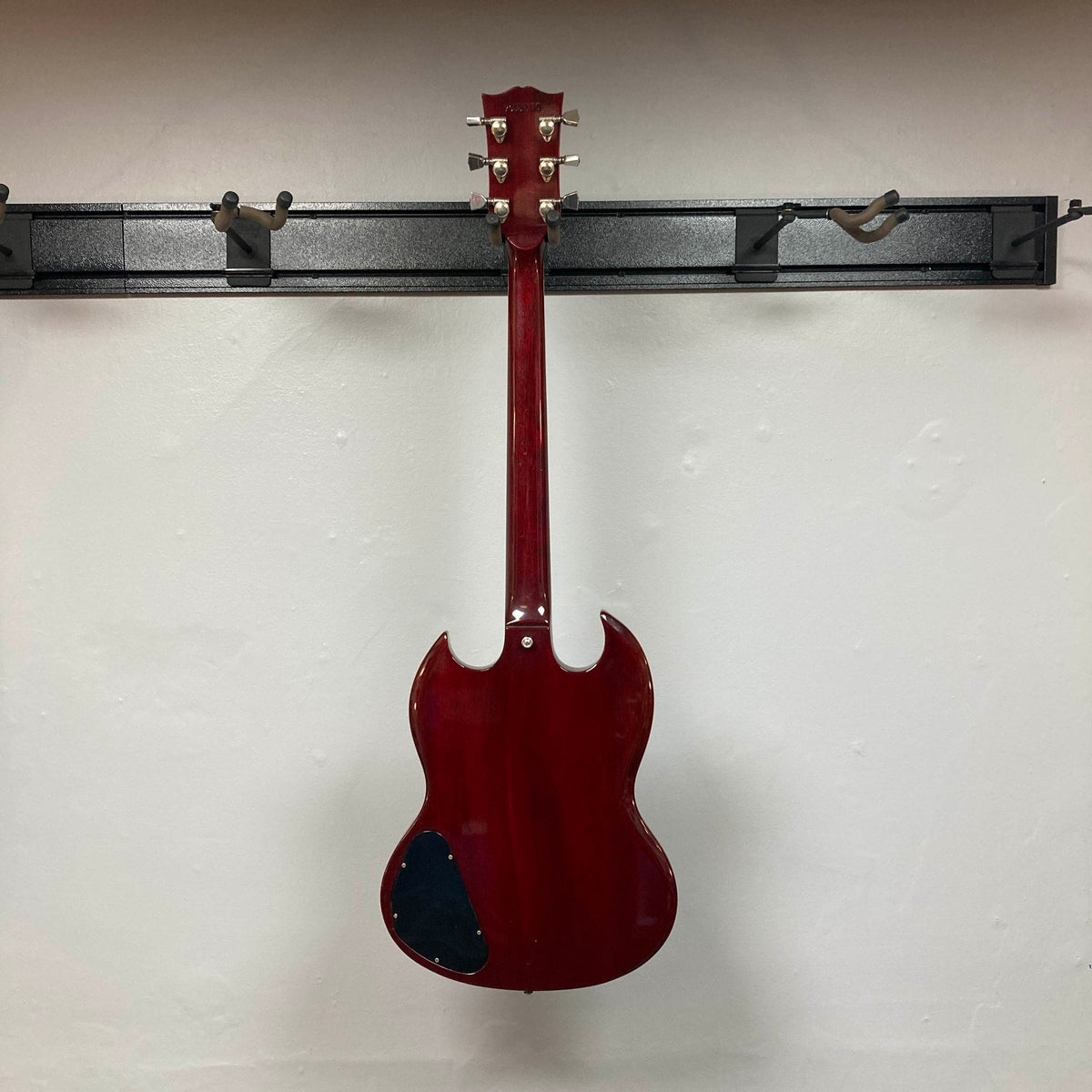 A 1977 Gibson SG Standard Cherry electric guitar by Gibson hangs on a wall-mounted rack, displayed from the back to reveal its visible tuning pegs and a black patch on the lower mahogany body.