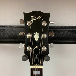 A close-up of the Gibson headstock showcases six tuning pegs, the iconic Gibson logo, and a distinctive inlay above a truss rod cover marked “S-G.” This 1977 Gibson SG Standard Cherry w/Case, made during the legendary Kalamazoo era, displays classic design elements that beautifully complement its sleek mahogany construction.