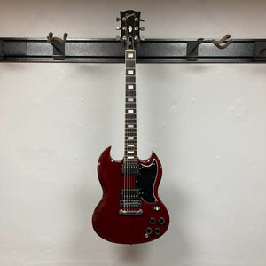 A 1977 Gibson SG Standard Cherry electric guitar by Gibson, featuring a mahogany body, black pickguard, and chrome hardware, hangs on a wall-mounted guitar holder against a white wall.