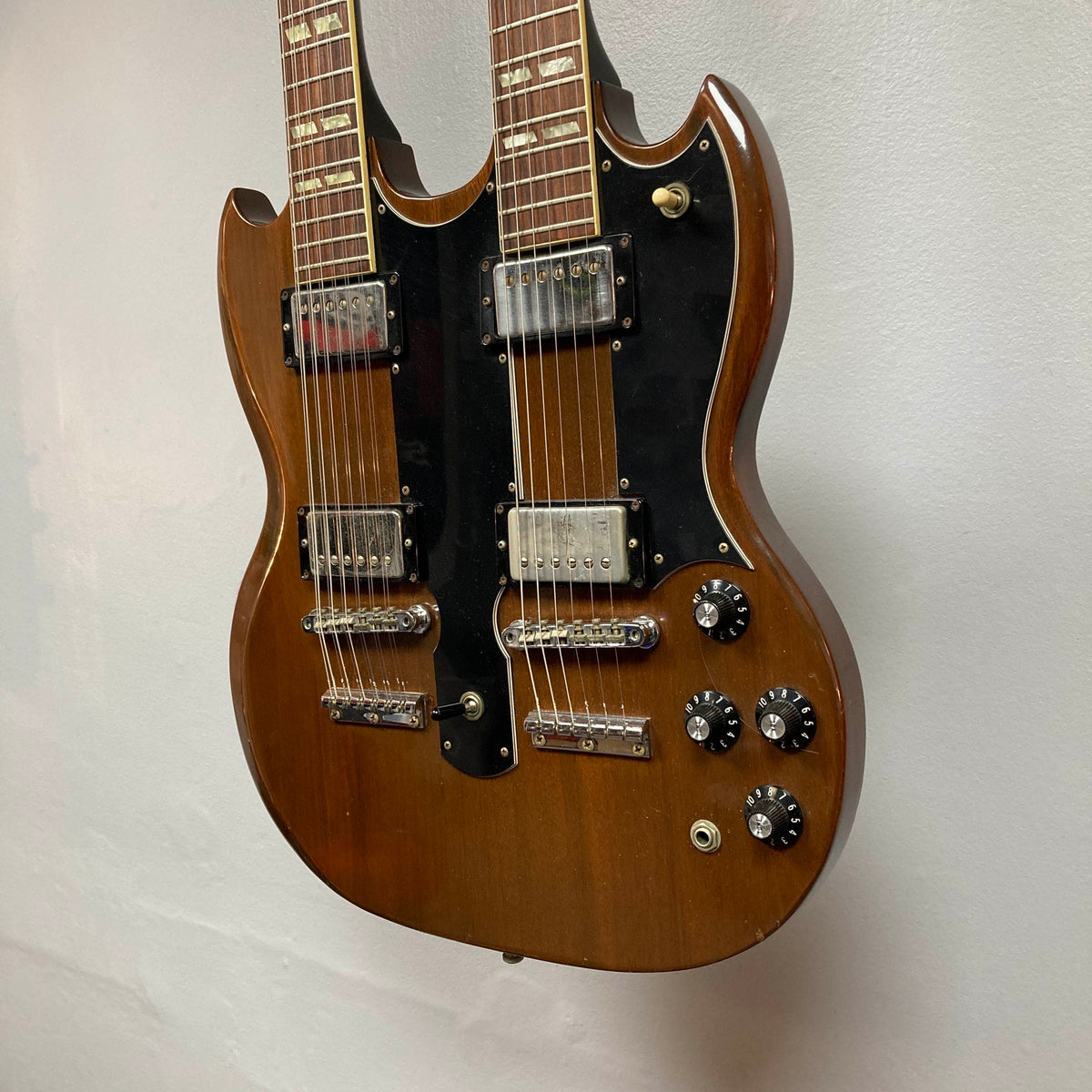 A 1978 Gibson EDS-1275 Double Neck Walnut w/Case by Gibson—featuring a rich brown walnut finish, black pickguard, four pickups, four control knobs, and metallic hardware—hangs against a plain wall, exemplifying style and versatility.