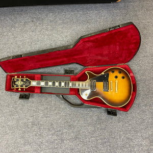A 1980 Gibson Les Paul Custom – Tobacco Sunburst w/OHSC by Gibson, featuring gold hardware, is displayed in its open, red velvet-lined original hard shell case on a gray carpeted floor—a vintage classic.