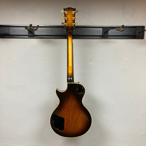 A 1980 Gibson Les Paul Custom – Tobacco Sunburst w/OHSC by Gibson hangs on a wall mount, photographed from the back with the control cavity covers removed, highlighting its vintage craftsmanship.