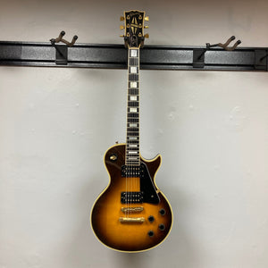 A 1980 Gibson Les Paul Custom – Tobacco Sunburst w/OHSC by Gibson, with gold hardware and black pickguard, hangs on a wall-mounted rack against a plain white wall, highlighting its classic vintage Les Paul style.