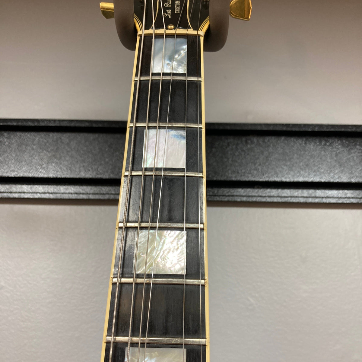 Close-up of a Gibson 1980 Les Paul Custom – Tobacco Sunburst w/OHSC neck, highlighting mother-of-pearl inlays, six strings, and gold tuners against a light gray wall—a striking vintage guitar from Gibson.