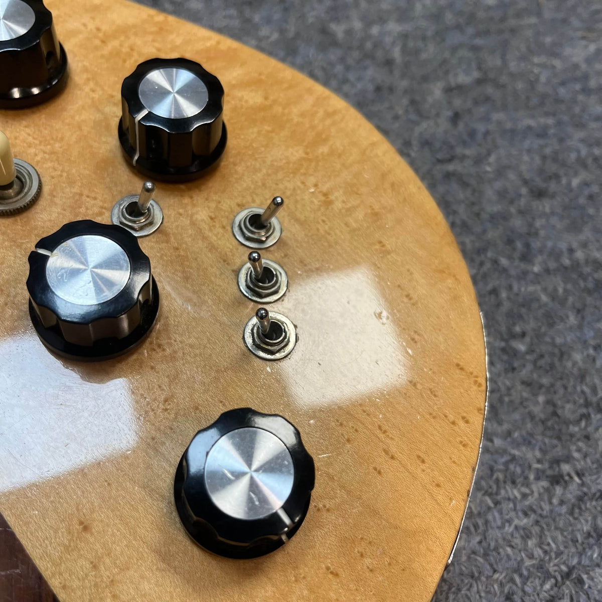 Close-up of a 1982 B.C. Rich Eagle Bass – USA Made with original electronics, three black knobs, and metal toggles on a light wood body with neck-through construction, professionally set up and shown on a gray textured surface.