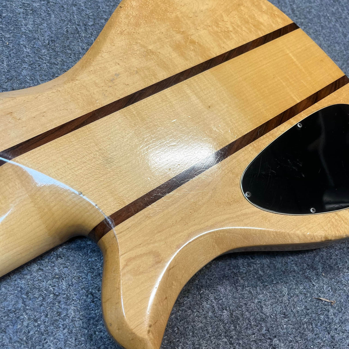 The back of a 1982 B.C. Rich Eagle Bass—USA made with original electronics—features dark wood stripes, a black plastic cover, and neck-through construction, highlighting its vintage appeal on a gray carpeted surface.