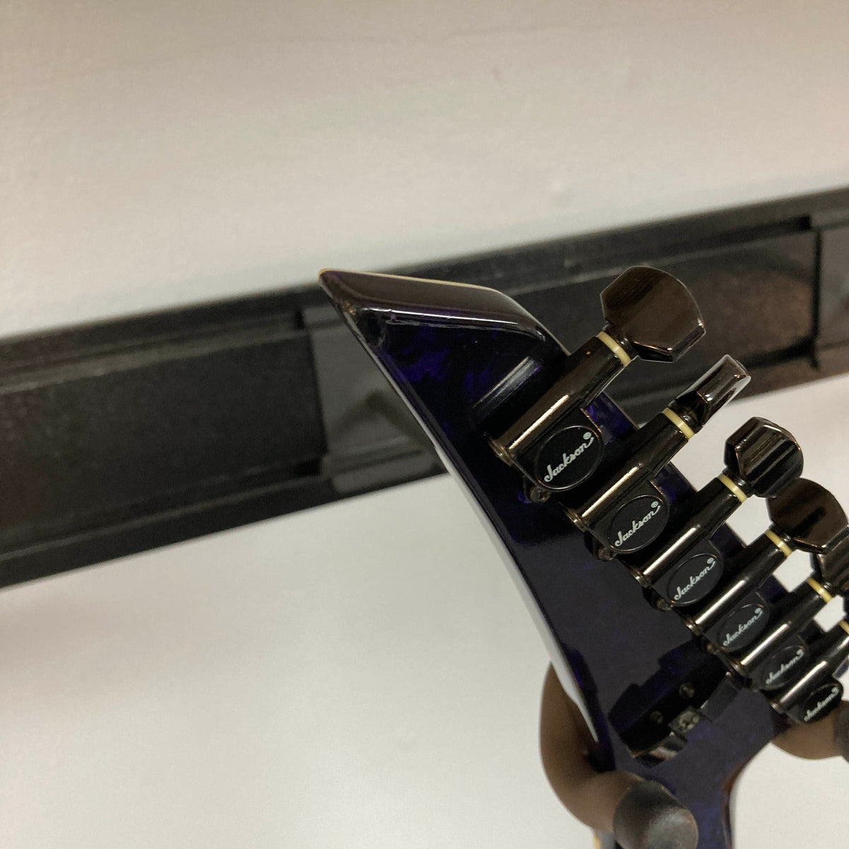 Close-up of the headstock of a Jackson 1995 SL1 Soloist Custom Shop Eerie Dess Swirl electric guitar, featuring black tuning pegs, mounted on a wall hanger against a white wall.