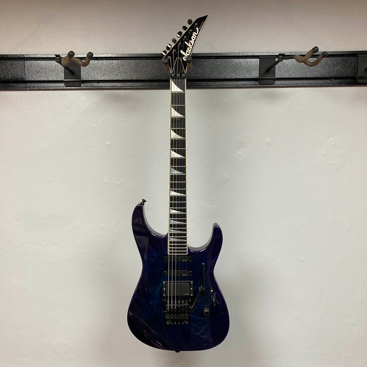 A 1995 Jackson SL1 Soloist Custom Shop Eerie Dess Swirl electric guitar by Jackson, featuring a pointed headstock and triangular inlays, is displayed on a wall-mounted guitar rack against a plain white wall.