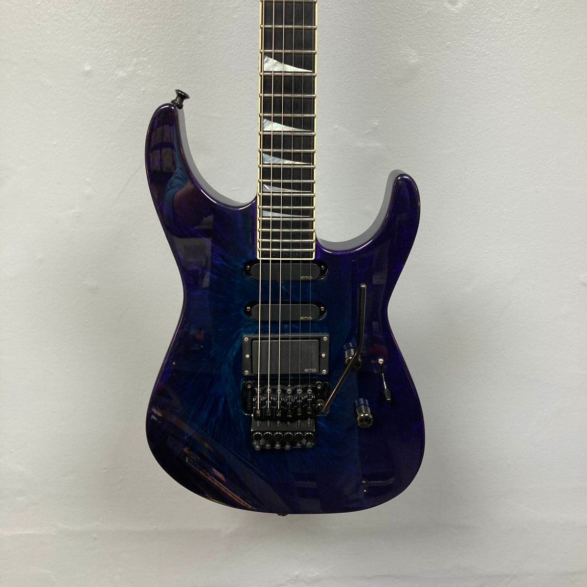 A 1995 Jackson SL1 Soloist Custom Shop Eerie Dess Swirl electric guitar by Jackson, featuring a striking blue double-cutaway body, three pickups, a Floyd Rose bridge, and sleek black hardware, hangs against a plain white wall.