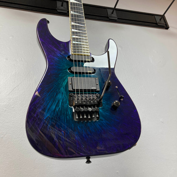 Hanging on the wall is a 1995 Jackson SL1 Soloist Custom Shop Eerie Dess Swirl electric guitar, featuring a striking purple and blue burst finish, black pickups, a Floyd Rose tremolo bridge, and a sleek maple neck—an iconic model from Jackson’s Custom Shop collection.