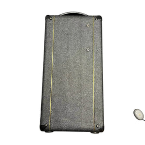 A rectangular, black textured Vox speaker cabinet with yellow lines stands upright; a small white wireless earbud rests in the corner, evoking the classic 2004 Vox AC30/6TB made in England with Celestion Alnico Blues.