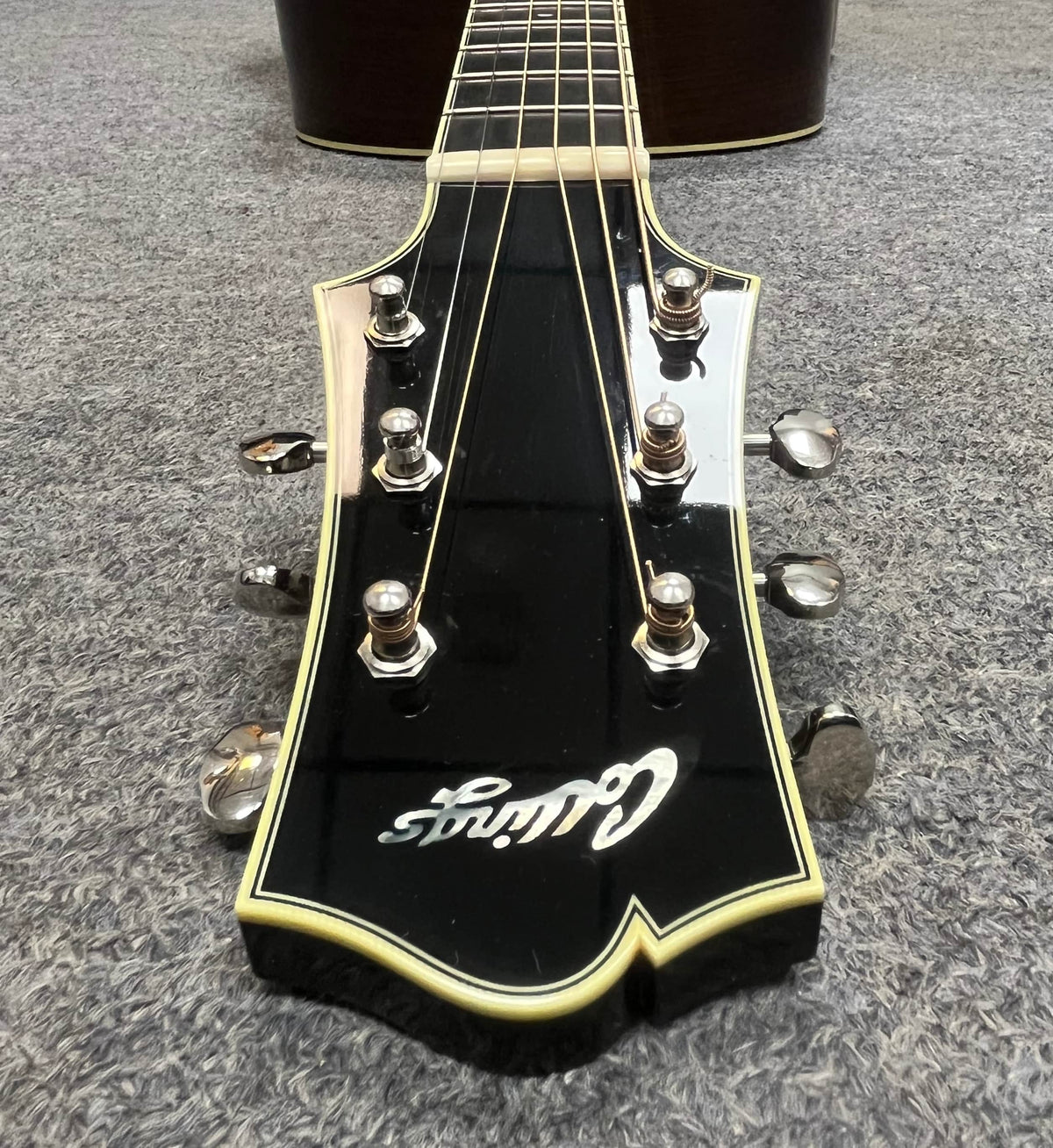 Close-up of a black headstock with six tuning pegs and cream binding, branded "Collings," from the 2005 Collings CJ BaA—Brazilian Rosewood & Adirondack Spruce—resting elegantly on a gray carpet.