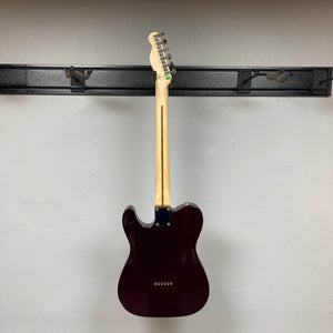 A 2007 Fender Standard Telecaster MIM in Midnight Wine Red by Fender hangs on a black wall rack, shown from the back. The light-colored neck and tuning pegs are visible; pickups and front details are not. The wall is plain and off-white.