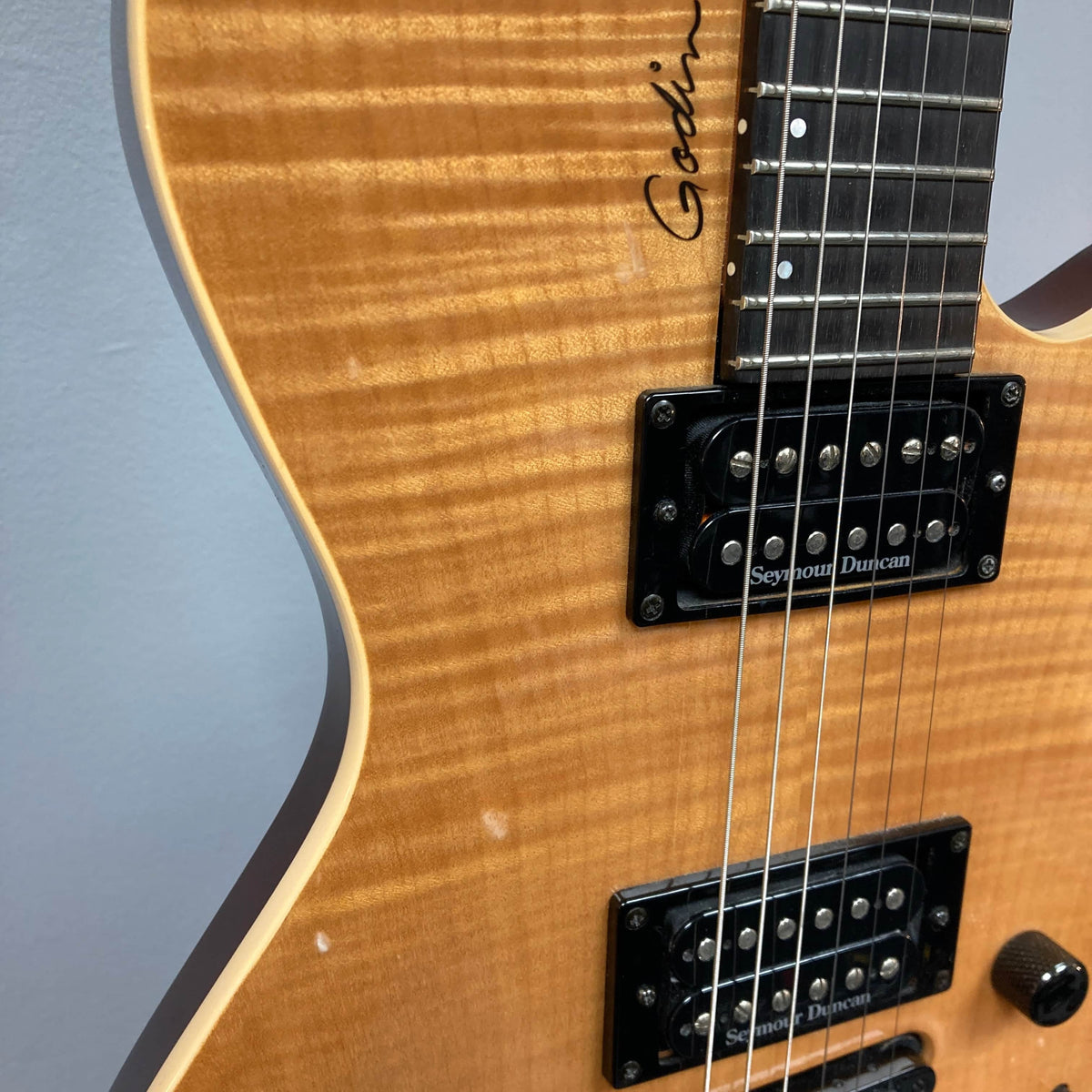 Close-up of a 2009 Godin Summit CT electric guitar with a carved flame maple cap, two humbuckers, part of the fretboard, and the Godin logo by the neck.