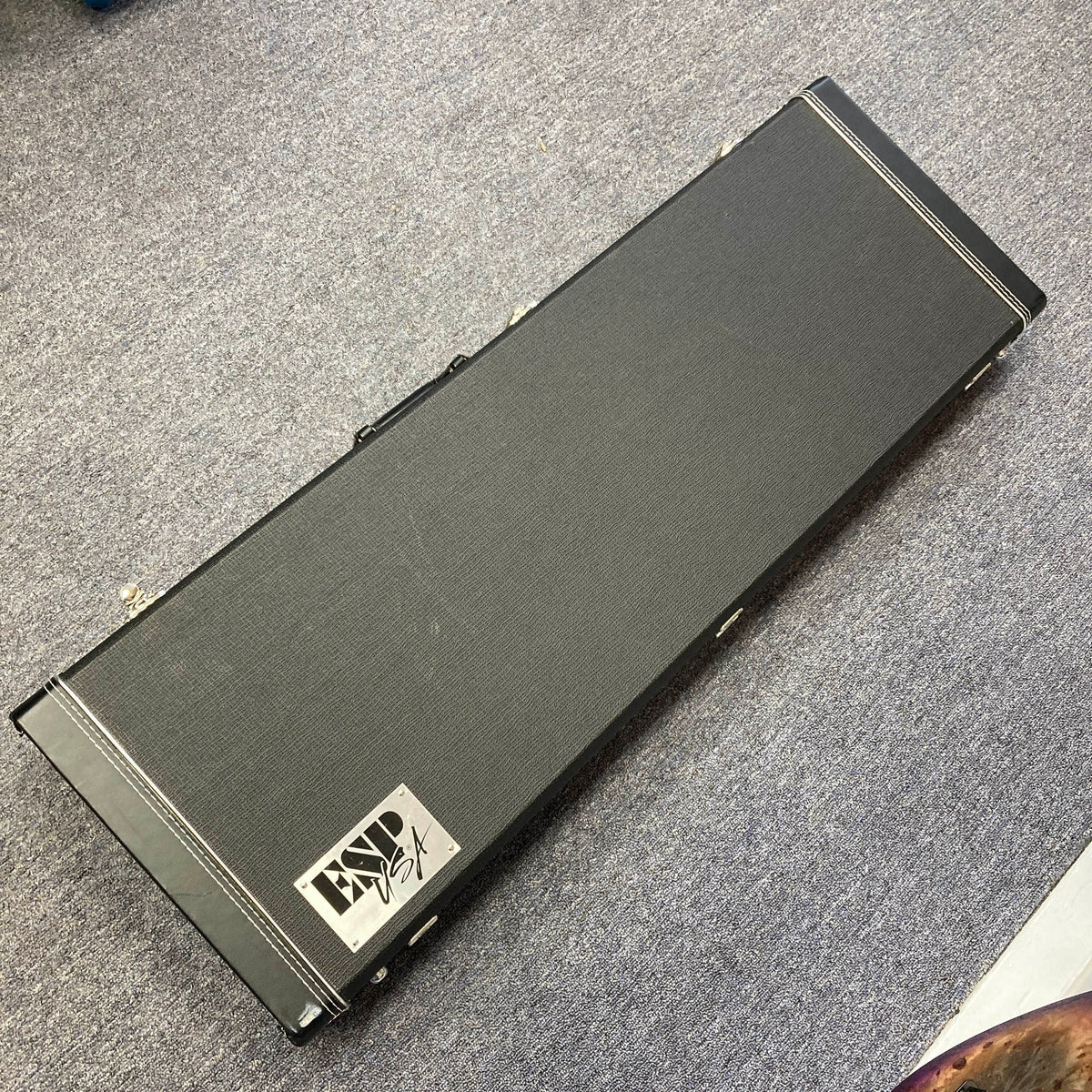 A black rectangular hard case with a handle, featuring a “Lynx USA” label, rests on a gray carpeted floor—perfect for safely storing your ESP 2019 USA M-II Purple Haze Burst electric guitar or proudly presenting this exceptional American-made ESP model.