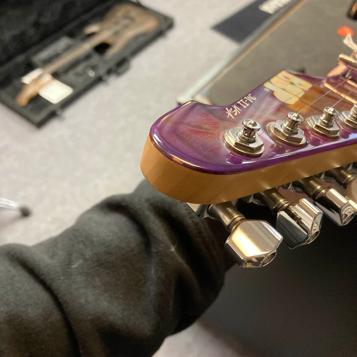 Close-up of the headstock and tuning pegs of a 2019 ESP USA M-II in striking Purple Haze Burst finish, highlighting one of ESP’s premier American-made guitars, with part of a person’s arm and the included case visible in the background.