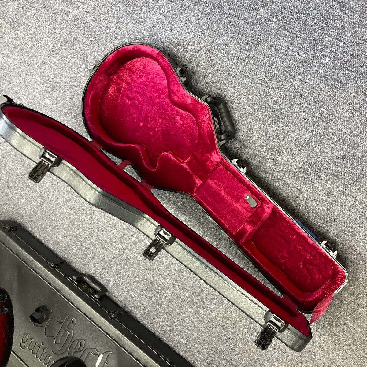 An open, empty Gator molded case with a red plush interior sits on a gray carpet, perfect for your 2025 Gibson Les Paul Studio Modern in Smokehouse Satin by Gibson. Another closed case rests nearby.