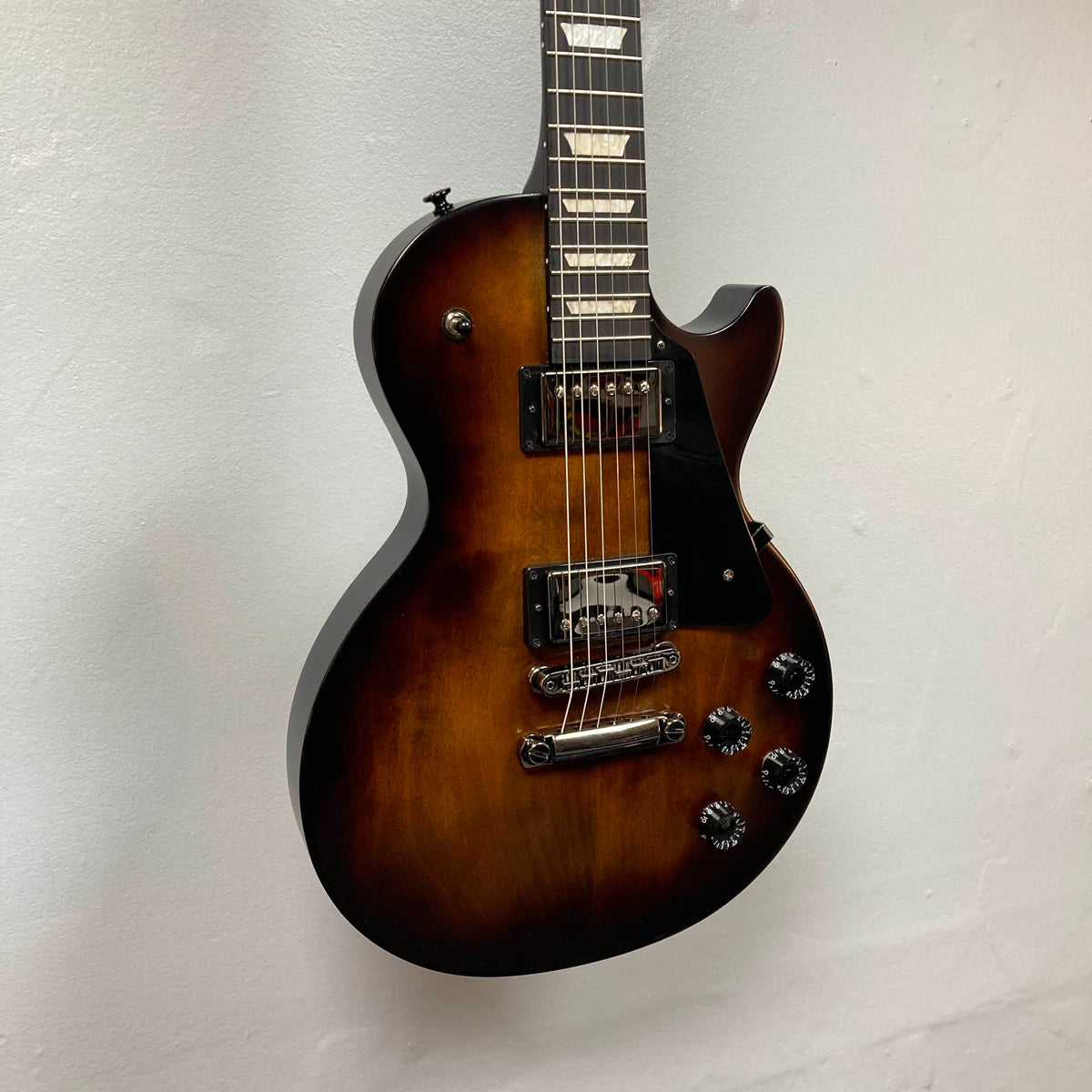 A 2025 Gibson Les Paul Studio Modern in Smokehouse Satin by Gibson, featuring chrome hardware and two humbuckers, hangs against a plain white wall. Comes with a Gator molded case.