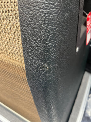 Close-up of the black textured surface of a 1969 Marshall 4x12 Cabinet with Matching Celestion T1281 Greenbacks, featuring a small scratch beside a brown woven panel reminiscent of classic Marshall styling, with part of a red sticker visible on the right.