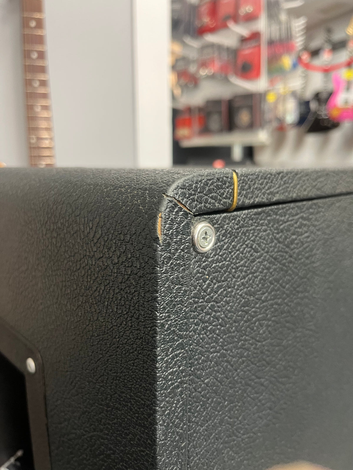 Close-up of a black textured corner on a 1969 Marshall 4x12 Cabinet with Matching Celestion T1281 Greenbacks, showing a tear in the tolex near a screw, with store shelves and a guitar neck blurred in the background.