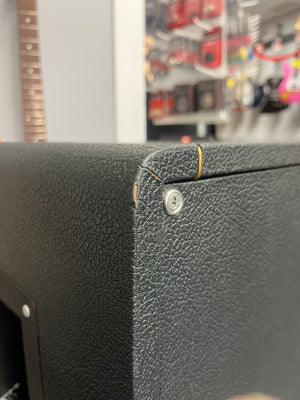 Close-up of a black textured corner on a 1969 Marshall 4x12 Cabinet with Matching Celestion T1281 Greenbacks, showing a tear in the tolex near a screw, with store shelves and a guitar neck blurred in the background.