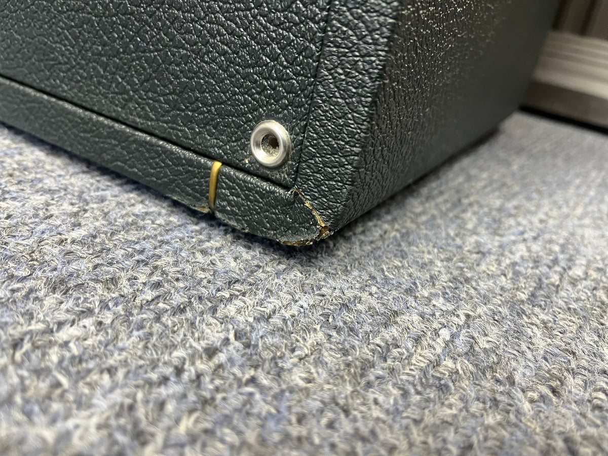 Close-up of the bottom corner of a textured black 1969 Marshall 4x12 Cabinet with Matching Celestion T1281 Greenbacks, featuring a small metal rivet, a rubber band, and a scuffed edge. The classic basketweave grille cloth, iconic to vintage Marshall cabinets, is visible as it rests on a gray carpeted floor.