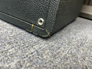 Close-up of the bottom corner of a textured black 1969 Marshall 4x12 Cabinet with Matching Celestion T1281 Greenbacks, featuring a small metal rivet, a rubber band, and a scuffed edge. The classic basketweave grille cloth, iconic to vintage Marshall cabinets, is visible as it rests on a gray carpeted floor.