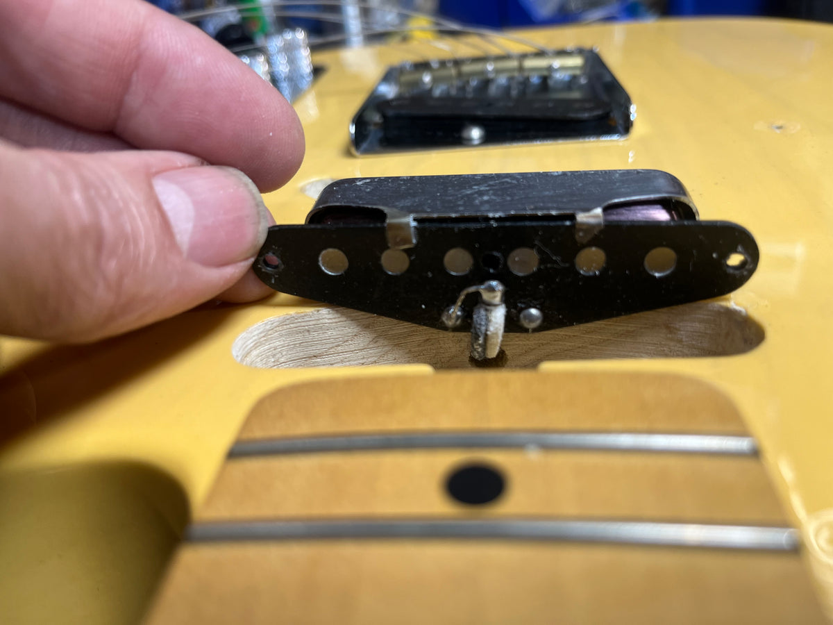 A hand holds a Fender Custom Shop '51 Reissue Nocaster NOS 2006 pickup above a yellow vintage blackguard guitar body, showing the wiring and cavity beneath.