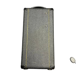 A rectangular, black textured Vox speaker cabinet with yellow lines stands upright; a small white wireless earbud rests in the corner, evoking the classic 2004 Vox AC30/6TB made in England with Celestion Alnico Blues.