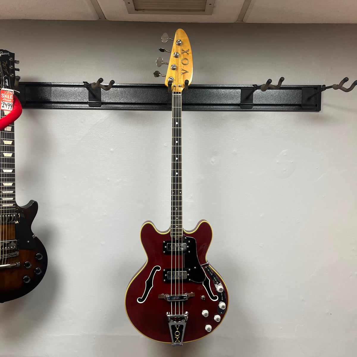 A red 1967-68 Vox Sidewinder IV Bass (V272) Cherry is hanging on a wall-mounted rack, its natural finish headstock visible next to a partially shown vintage Italian bass.