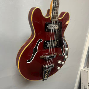 A 1967-68 Vox Sidewinder IV Bass (V272) in cherry by Vox, a semi-hollow electric bass with white trim, two humbuckers, four control knobs, and f-shaped sound holes—vintage Italian style—hangs on the wall.