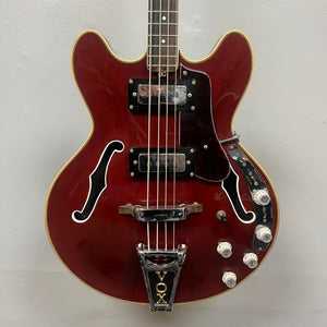 The 1967-68 Vox Sidewinder IV Bass (V272) Cherry by Vox is a red semi-hollow electric bass with two pickups, four white control knobs, and a VOX-marked Bigsby-style tremolo, echoing the classic vintage Italian design.