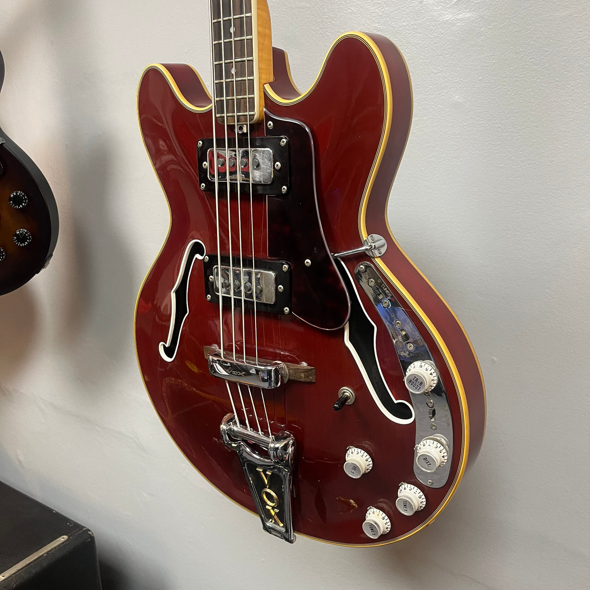 A 1967-68 Vox Sidewinder IV Bass (V272) Cherry by Vox, this semi-hollow red electric bass with white trim, multiple control knobs, and two pickups hangs on the wall—evoking classic vintage style.