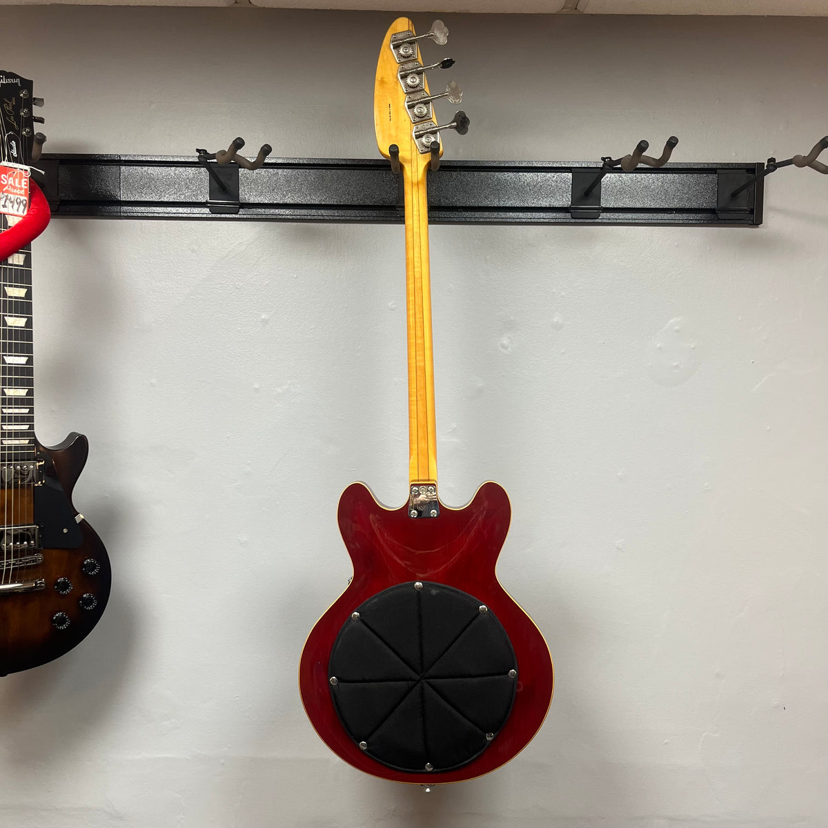 A cherry 1967-68 Vox Sidewinder IV Bass (V272) by Vox, with a circular black practice pad attached, hangs on a wall-mounted rack next to a dark electric guitar.