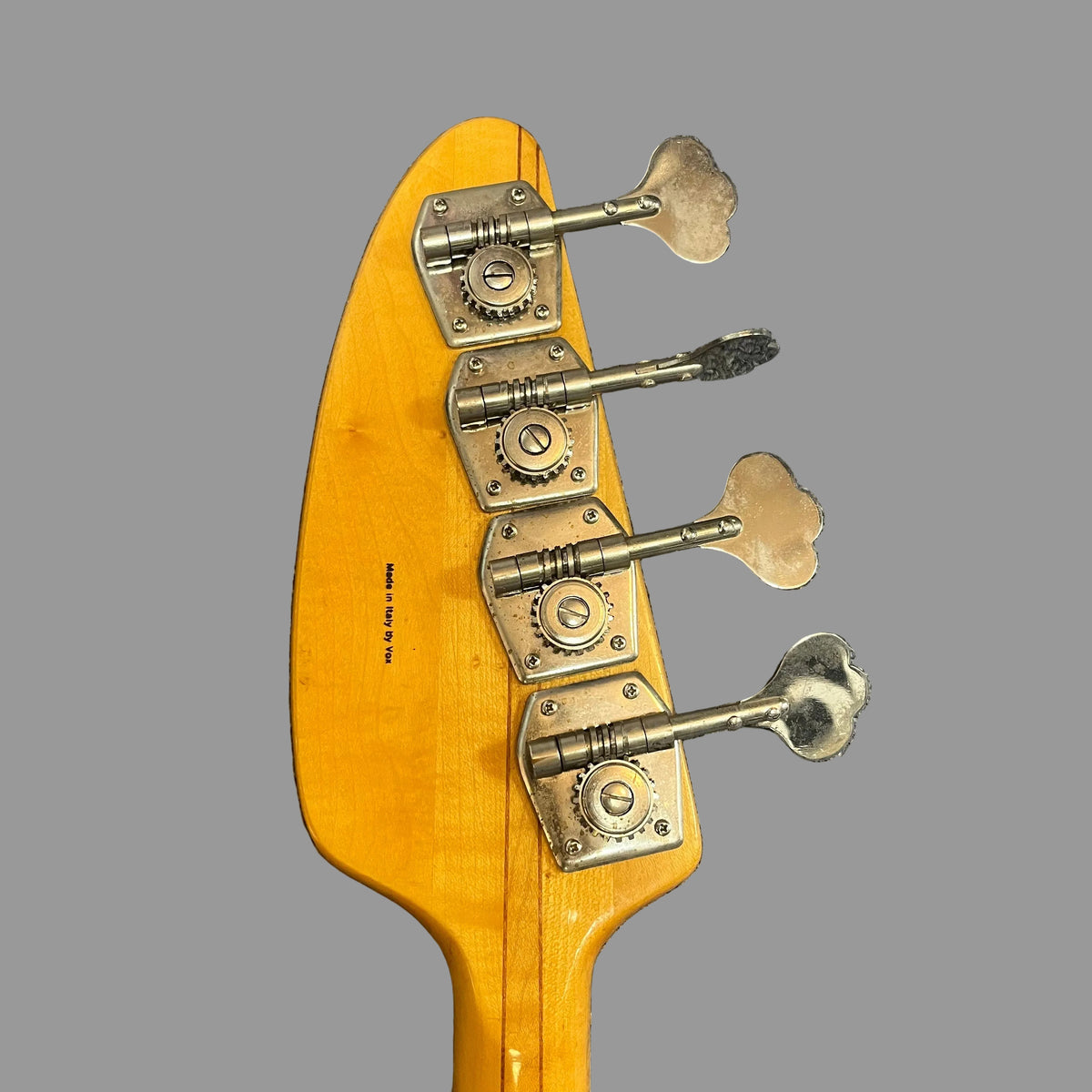 Close-up of the back of a 1967-68 Vox Sidewinder IV Bass (V272) Cherry headstock by Vox, featuring four silver tuning pegs against a plain gray background.