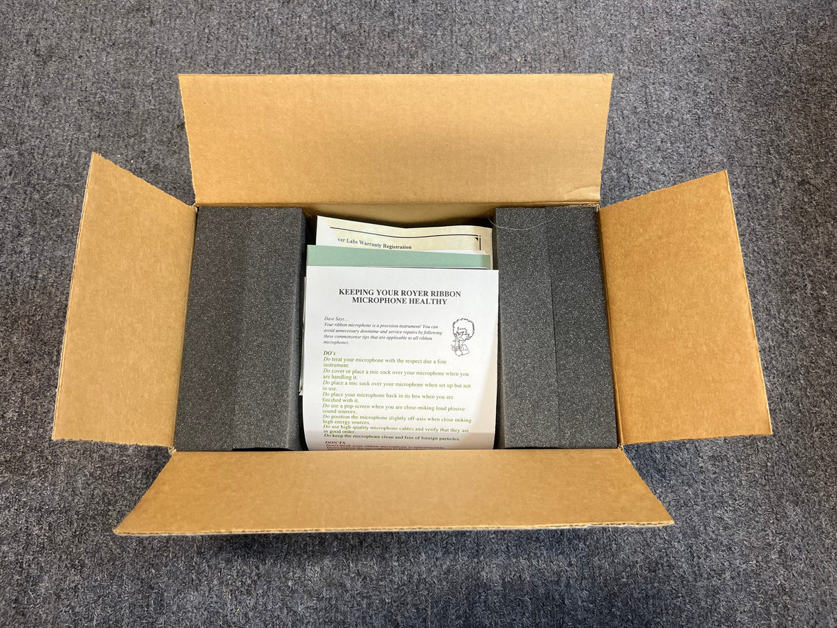 An open cardboard box on a gray carpet holds documents and foam padding, with an instruction sheet on top—ideal for anyone using the Royer Labs R-121 Ribbon Microphone (Pre-owned in excellent condition) by Royer Labs.