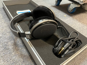 A pair of preowned Sennheiser HD 650 Reference Class headphones, featuring an open-back black design with a coiled cable and adapter, are displayed in a foam-lined case on a grey carpeted surface.