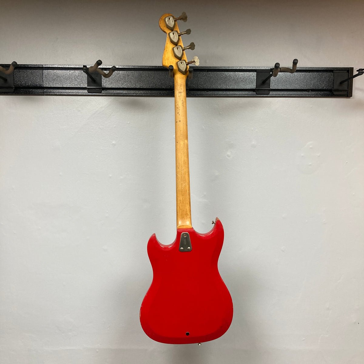 Hagstrom HII-B Mid-1960s Red Medium-Scale Bass, serial 643772, hanging on a wall rack, showcasing its double-cutaway design and maple neck with four tuning pegs.
