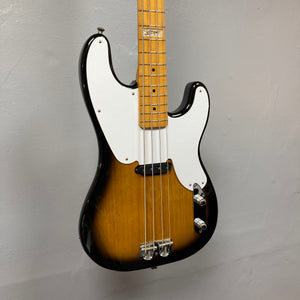 A close-up of the Fender Sting P Bass Sunburst, a vintage-style electric bass by Fender with a sunburst finish, white pickguard, and four strings, hanging on a light gray wall.