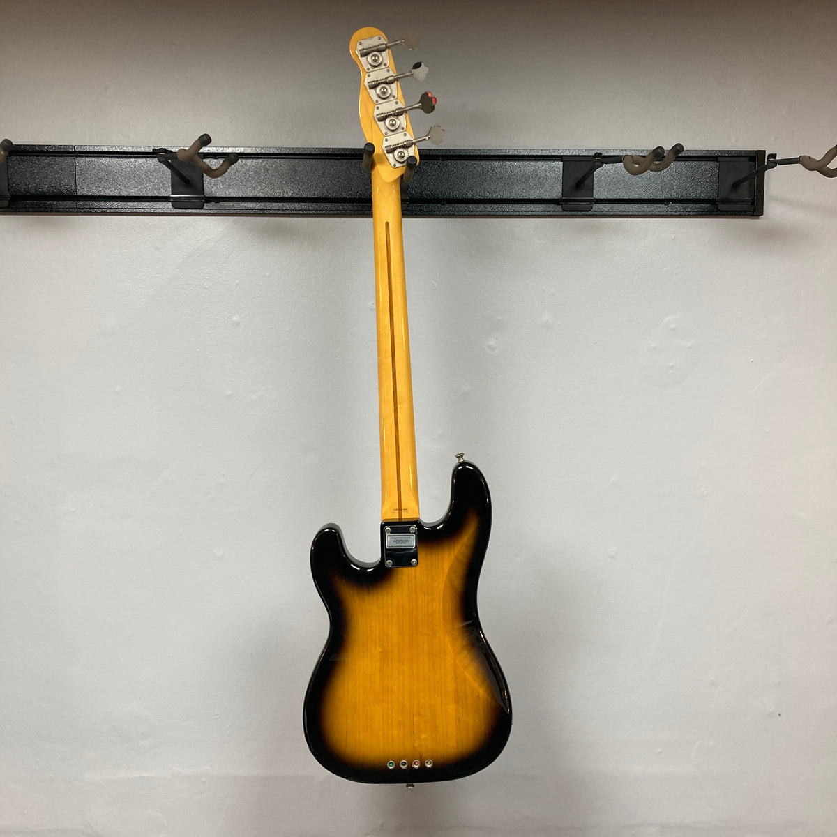 A Fender Sting P Bass Sunburst by Fender hangs from its neck on a black wall-mounted rack against a plain white wall.