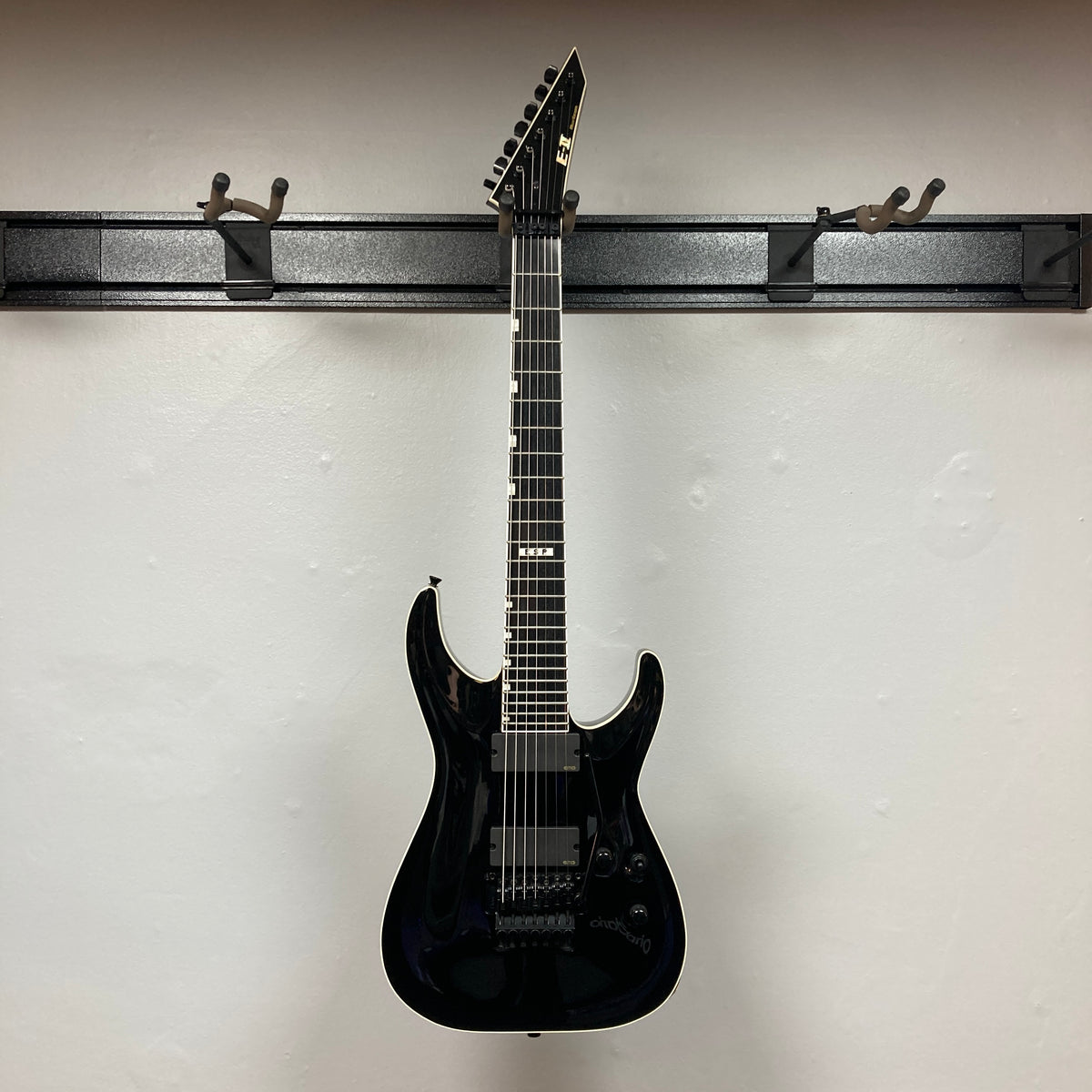 An ESP E-II Horizon FR-7 Black w/Case 7-string electric guitar by ESP with a pointed headstock and Floyd Rose bridge hangs on a wall-mounted rack against a plain white wall.