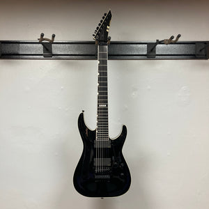 An ESP E-II Horizon FR-7 Black w/Case 7-string electric guitar by ESP with a pointed headstock and Floyd Rose bridge hangs on a wall-mounted rack against a plain white wall.