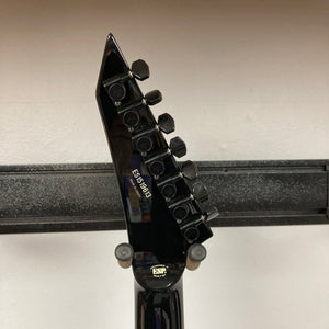 Close-up of the ESP E-II Horizon FR-7 Black electric guitar headstock, showing six tuning pegs and serial number ES176969, resting on a wall-mounted holder. Comes with case. Brand: ESP.